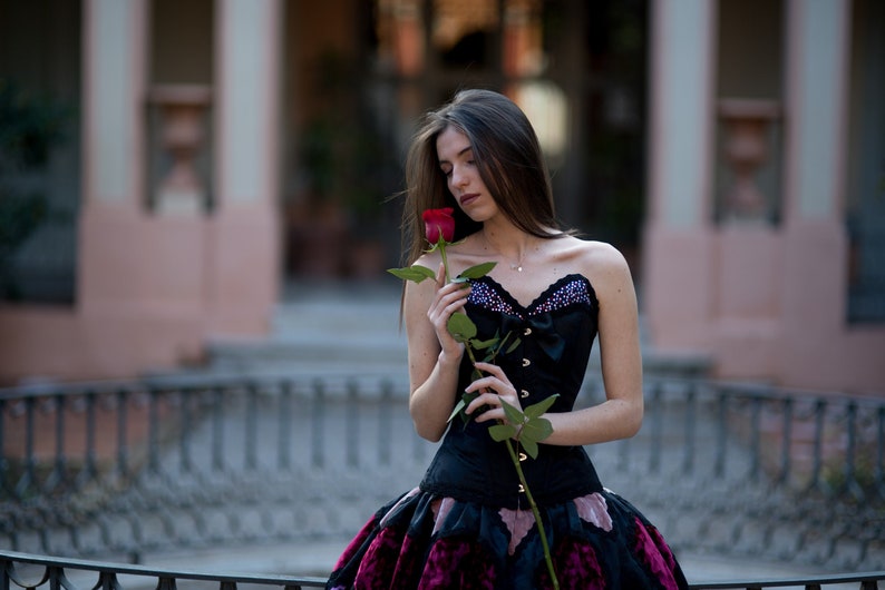 K&ouml;nnte beinhalten: Eine Frau in einem schwarzen Korsett und einem rosa-schwarzen Rock h&auml;lt eine einzelne rote Rose. Sie steht vor einem Geb&auml;ude mit einem schmiedeeisernen Zaun im Vordergrund.