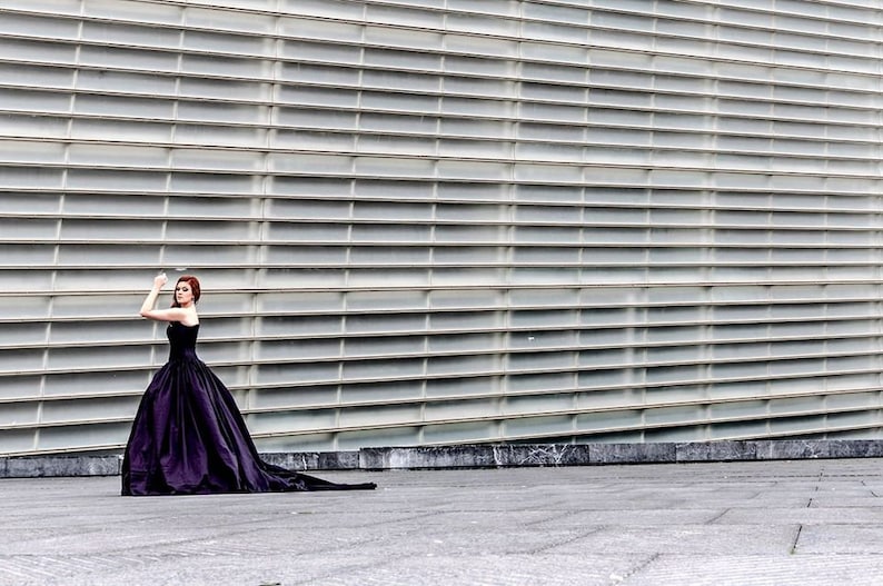 May include: A floor-length, strapless black ball gown with a fitted bodice and a voluminous, full skirt that extends into a long train. The garment features a smooth, solid-colored fabric and a structured silhouette that flares from the waist down.