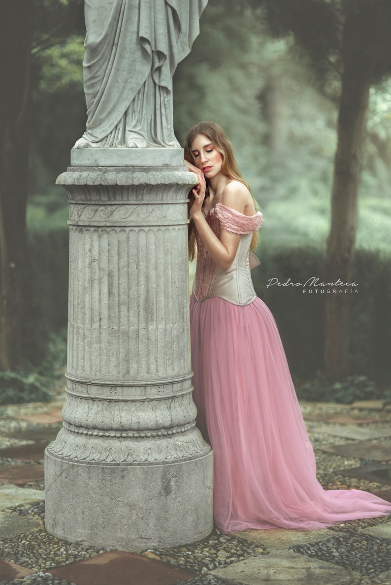 May include: A woman in a pink tulle dress leans against a stone pillar in a garden setting. The pillar is decorated with intricate carvings. The woman's dress is flowing and elegant, and her hair is long and flowing. The background is blurred, creating a dreamy atmosphere.