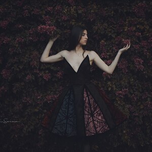May include: A woman in a black dress with a colorful patterned skirt stands in front of a wall of purple flowers.