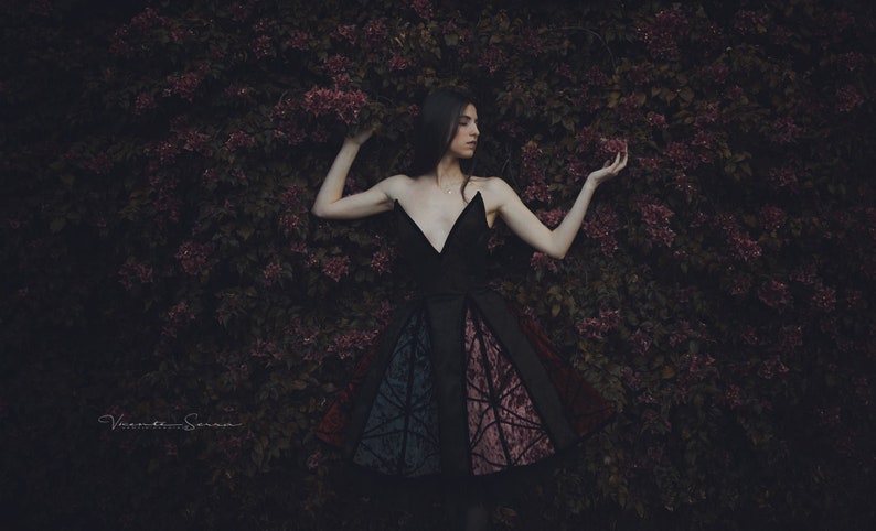May include: A woman in a black dress with a colorful patterned skirt stands in front of a wall of purple flowers.