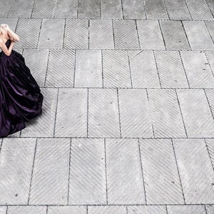 May include: A floor-length, strapless deep purple gown with a fitted bodice and a voluminous, pleated skirt that flares out into a wide train, featuring a smooth, satin-like fabric with a subtle sheen that catches the light across the folds.
