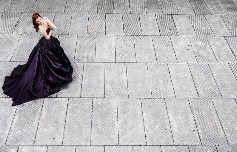 May include: A floor-length, strapless deep purple gown with a fitted bodice and a voluminous, pleated skirt that flares out into a wide train, featuring a smooth, satin-like fabric with a subtle sheen that catches the light across the folds.