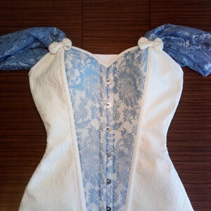 May include: A white and blue corset with lace detailing and a busk closure. The corset has a sweetheart neckline and is made of a textured fabric.