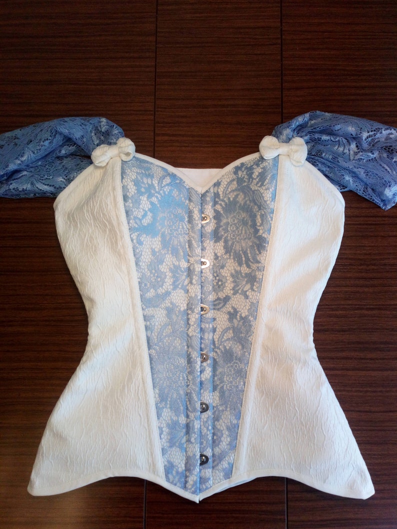 May include: A white and blue corset with lace detailing and a busk closure. The corset has a sweetheart neckline and is made of a textured fabric.