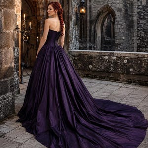 May include: A floor-length, strapless deep purple gown featuring a fitted bodice with a lace-up back closure and a voluminous, pleated skirt that extends into a long train. The fabric has a subtle sheen and a structured, full silhouette.