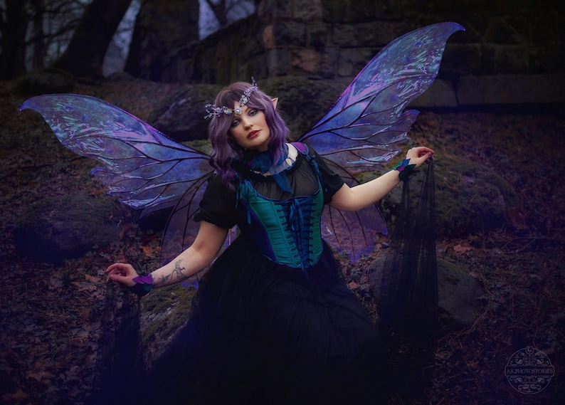 May include: A woman with purple hair and iridescent wings is dressed in a black dress with a teal corset. She is wearing a crown of flowers and has a dark background.