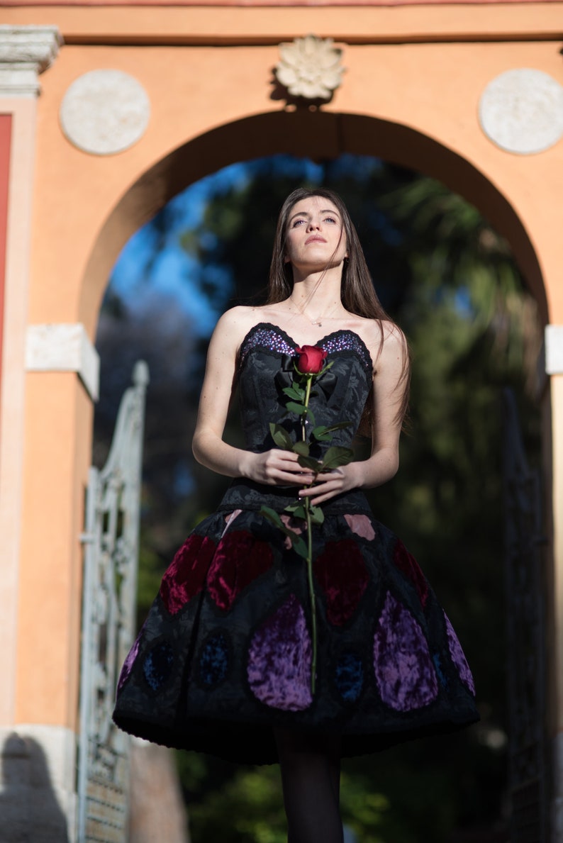 K&ouml;nnte beinhalten: Eine Frau in einem schwarzen Kleid mit bunten Samtdetails h&auml;lt eine einzelne rote Rose. Sie steht vor einem Torbogen mit einem Steinbogen und gr&uuml;nem Hintergrund.