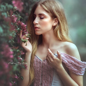 May include: A woman with long blonde hair wearing a light blue and pink corset top with lace details. She is standing in front of a bush of pink flowers and looking down at the flowers.