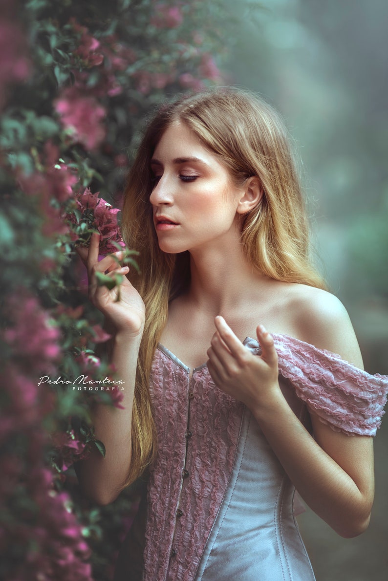 May include: A woman with long blonde hair wearing a light blue and pink corset top with lace details. She is standing in front of a bush of pink flowers and looking down at the flowers.