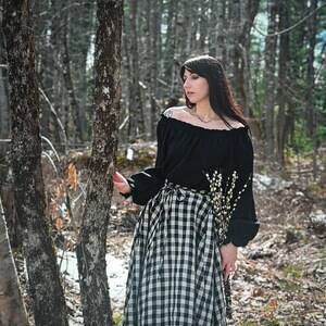 May include: A woman wearing a black off-the-shoulder blouse and a black and white gingham skirt stands in a forest. She is holding a bouquet of white flowers.