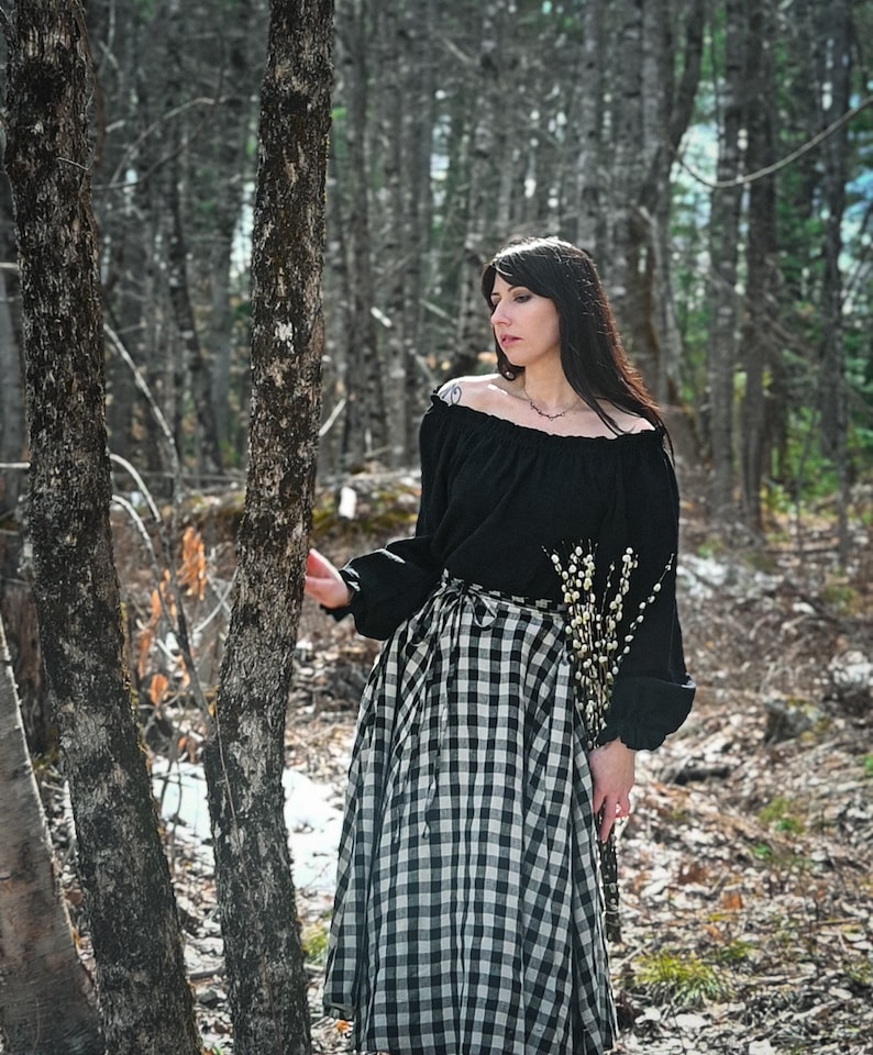 May include: A woman wearing a black off-the-shoulder blouse and a black and white gingham skirt stands in a forest. She is holding a bouquet of white flowers.