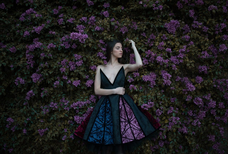 May include: A woman in a black, blue, and purple dress with a geometric pattern stands in front of a wall of purple flowers.