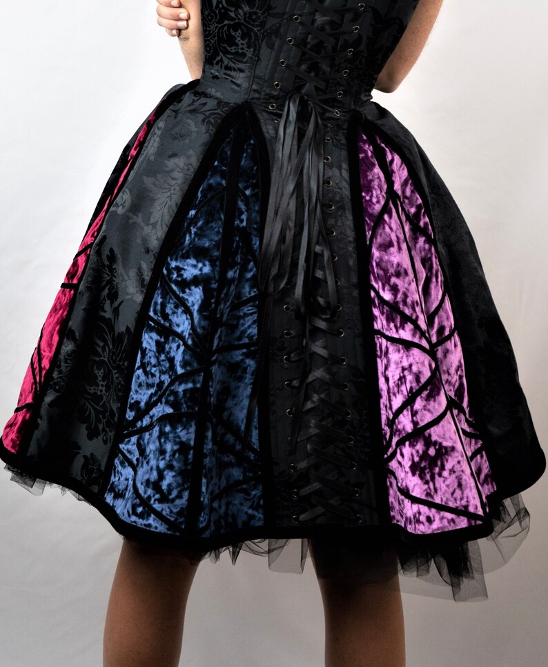May include: A black, velvet, and satin dress with a fitted bodice and a full skirt. The skirt is made of four panels of different colors: red, blue, black, and purple. The dress has a lace-up back.