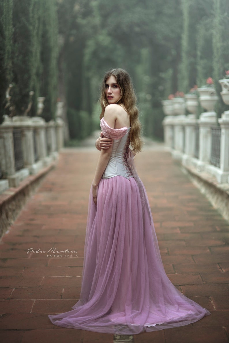 May include: A woman in a white corset and a long, flowing pink tulle skirt stands on a brick pathway. The background is a blurred image of a garden with trees and bushes.