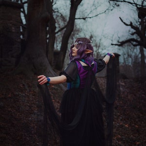 May include: A woman with purple hair and pointed ears wears a black dress with a purple and teal bodice. She is standing in a forest with trees in the background. She is holding the edges of her dress and looking to the side.