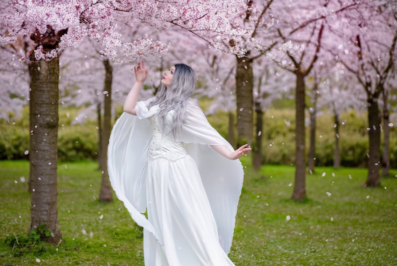 May include: A woman wearing a white dress and cape stands in a grove of cherry blossom trees. The trees are in full bloom and petals are falling around her.