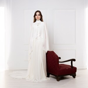 May include: A woman in a white gown stands in a white room with a red armchair. The gown has a high neck and long sleeves. The woman has long brown hair and is looking at the camera.