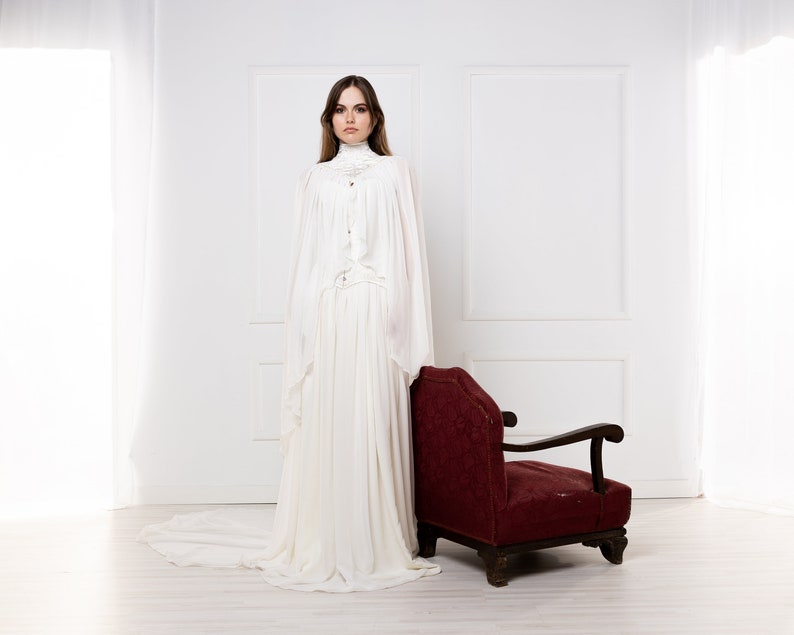 May include: A woman in a white gown stands in a white room with a red armchair. The gown has a high neck and long sleeves. The woman has long brown hair and is looking at the camera.
