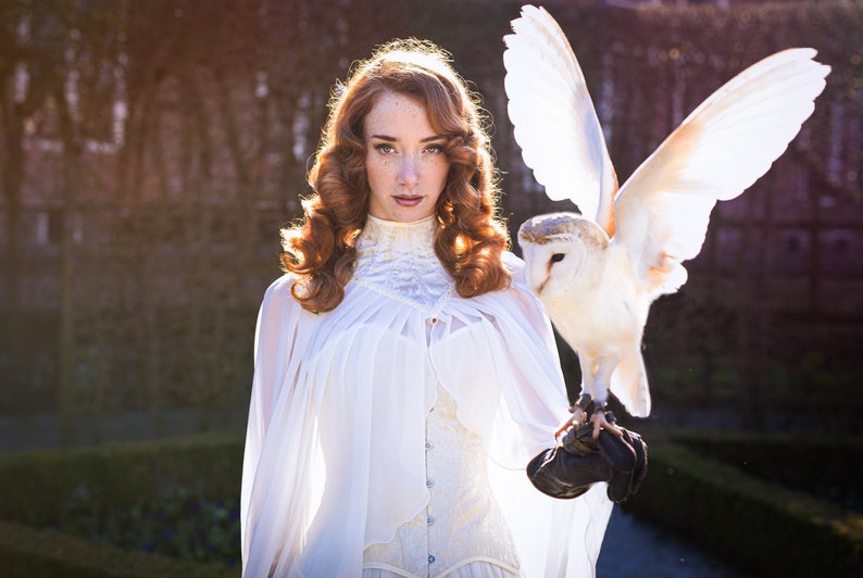May include: A woman with long, curly red hair wearing a white dress and a white cape holds a white owl with its wings spread out. The owl is perched on a gloved hand.