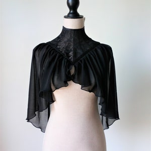 May include: A black sheer cape with a lace collar and ruffled edges. The cape is displayed on a white mannequin.