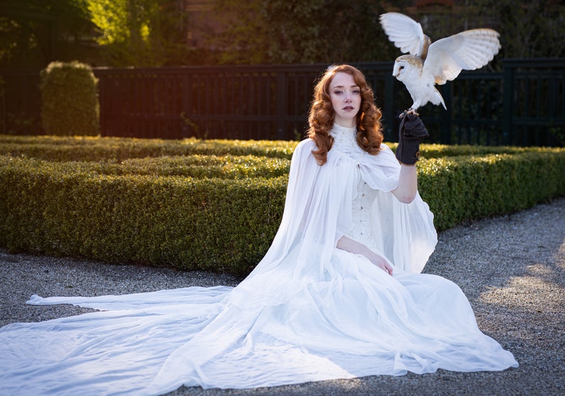 May include: A woman wearing a white gown and cape sits in a garden with a white owl perched on her gloved hand.