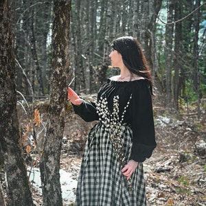 May include: A woman in a black off-the-shoulder blouse and a black and white gingham skirt stands in a forest. She is holding a bouquet of white pussy willow branches.