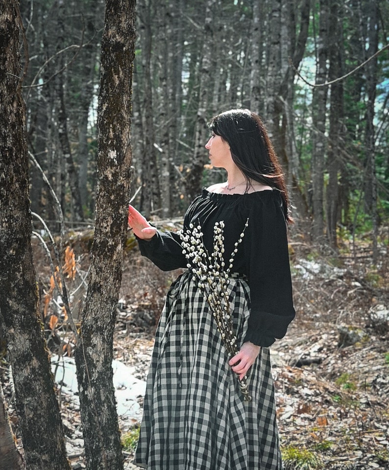 May include: A woman in a black off-the-shoulder blouse and a black and white gingham skirt stands in a forest. She is holding a bouquet of white pussy willow branches.