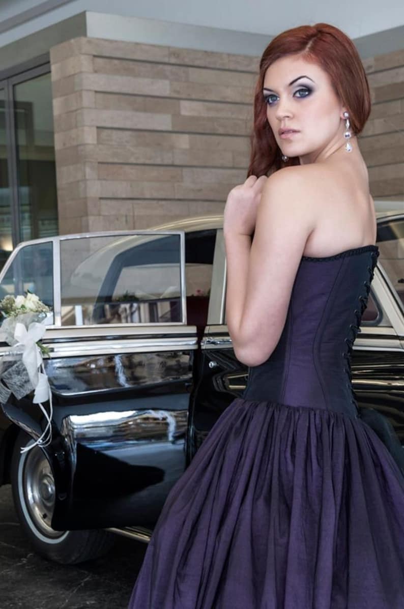 May include: A strapless dark purple corset dress featuring a fitted bodice with black lace-up detailing on the back and a full, voluminous skirt. The garment is worn with long, dangling silver earrings with clear teardrop-shaped stones.