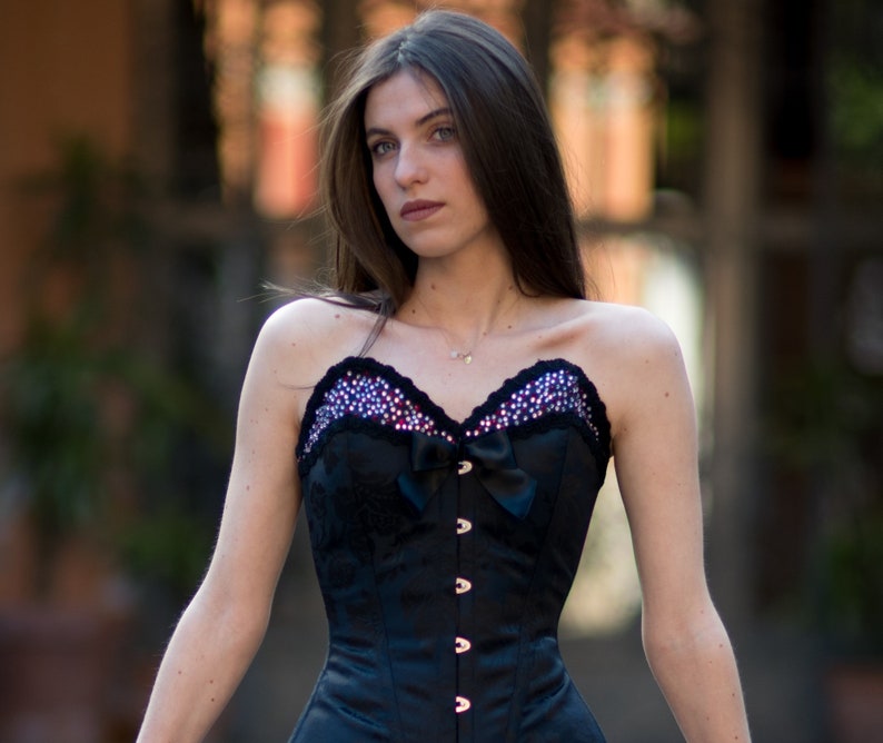 K&ouml;nnte beinhalten: Ein schwarzes, tr&auml;gerloses Korsett mit Satin-Finish und einer Spitzenborte. Das Korsett hat eine Schleife in der Mitte und ist mit Strasssteinen verziert.