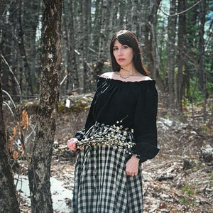 May include: A woman in a black off-the-shoulder blouse and a black and white checkered skirt stands in a forest. She is holding a bouquet of white flowers.