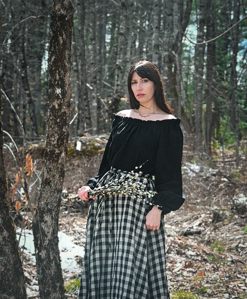 May include: A woman in a black off-the-shoulder blouse and a black and white checkered skirt stands in a forest. She is holding a bouquet of white flowers.