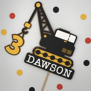 May include: A black and yellow construction crane cake topper with the number 3 and the name Dawson. The crane is holding a hook with a yellow circle on it. The topper is on a wooden stick.