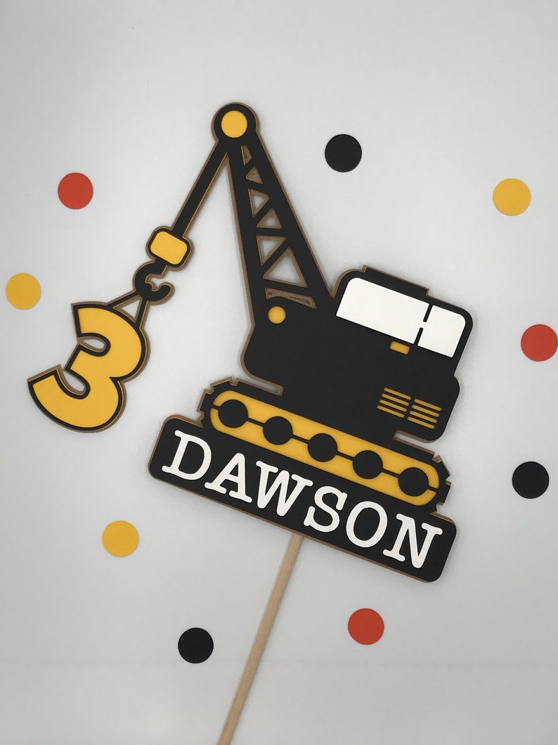 May include: A black and yellow construction crane cake topper with the number 3 and the name Dawson. The crane is holding a hook with a yellow circle on it. The topper is on a wooden stick.