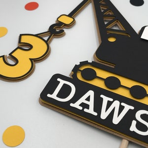 May include: A yellow and black construction crane cake topper with the number 3 and the name "DAWS" on it.