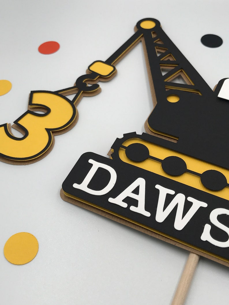 May include: A yellow and black construction crane cake topper with the number 3 and the name "DAWS" on it.