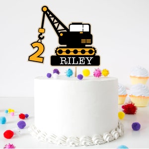 May include: A yellow and black construction crane cake topper with the number 2 and the name Riley. The topper is on a white cake with colorful pom-poms around it.
