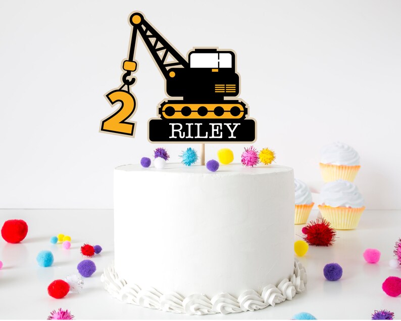 May include: A yellow and black construction crane cake topper with the number 2 and the name Riley. The topper is on a white cake with colorful pom-poms around it.