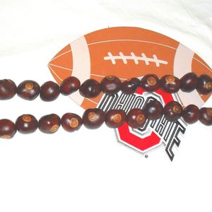 May include: A necklace made of dark brown buckeye nuts, displayed with a football-shaped cutout and an Ohio State logo. The buckeyes are strung together, creating a unique and natural accessory. The background is white.