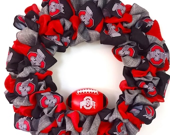 May include: A circular wreath made of red, black, and grey ribbons, adorned with the Ohio State logo. A red football, also bearing the Ohio State logo, is positioned in the centre. Ideal for door or wall decoration.