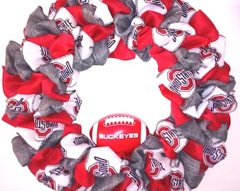 May include: A circular fabric wreath featuring a repeating pattern of red, white, and gray Ohio State University logos and text. A small red and white football with the word BUCKEYES printed in white is attached to the center of the wreath.