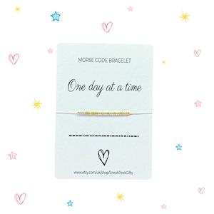May include: A Morse code bracelet with gold and silver beads, strung on a fine cord. The accompanying card says "One day at a time". The background is white with decorative hearts and stars in pink and yellow.