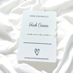 May include: A white card with the text "Morse Code Bracelet" and "Fuck Cancer" in black cursive font. Below the text is a string bracelet with white and gold beads. The bracelet is tied in a knot. Below the bracelet is a line of dots and dashes representing the Morse code for "Fuck Cancer". Below the Morse code is a heart symbol. The card also includes the website address "www.etsy.com/uk/shop/SneakPeekGifts".