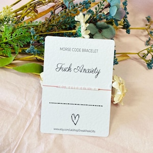 May include: A white card with the text "Morse Code Bracelet" and "Fuck Anxiety" in cursive. A pink string bracelet with white beads is displayed on the card. The Morse code for the phrase is shown below the bracelet. The card also includes a heart symbol and the website address "www.etsy.com/uk/shop/SneakPeekGifts".