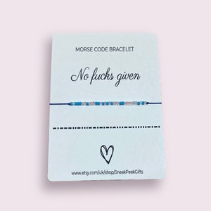 May include: A white card with the text "Morse Code Bracelet" and "No fucks given" in black cursive font. The card shows a blue and white beaded bracelet with a black string. The card also includes a heart symbol and the website address "www.etsy.com/uk/shop/SneakPeekGifts".