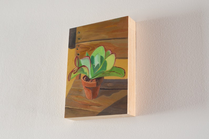Puede incluir: Pintura de una planta suculenta en una maceta de terracota. La planta tiene hojas verdes con bordes rojos. La maceta est&aacute; sobre una superficie de madera, con un fondo amarillo c&aacute;lido. La obra de arte est&aacute; sobre un panel de madera.