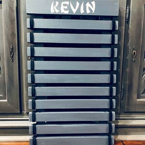 Może przedstawiać: Drewniany stojak na pasy w kolorze szarym z czarnymi paskami i napisem "KEVIN" w kolorze białym na g&oacute;rze.