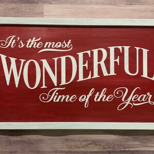 Puede incluir: Un letrero de madera rojo con marco blanco y el texto "It's the most WONDERFUL Time of the Year" en letras blancas cursivas.