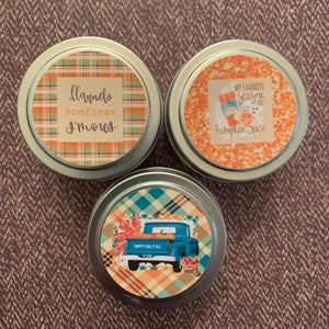 May include: Three round metal tins with colourful labels. The first tin has a plaid pattern and the text "flannels bonfires s'mores". The second tin has a pumpkin spice latte graphic and the text "My favourite season is pumpkin spice". The third tin has a plaid pattern and a blue pickup truck with the text "Happy Fall Y'all".