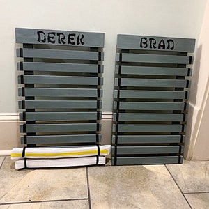 Może przedstawiać: Dwa szare drewniane stojaki na pasy z czarną ozdobą i nadrukowanymi nazwami "Derek" i "Brad". Biały i ż&oacute;łty pas leży na podłodze przed stojakiem.